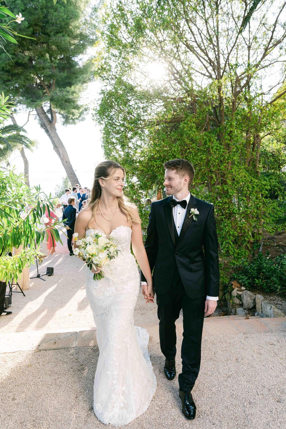 Bride in lace gown and groom in black tuxedo hold hands walking through a sunlit garden on their wedding day.