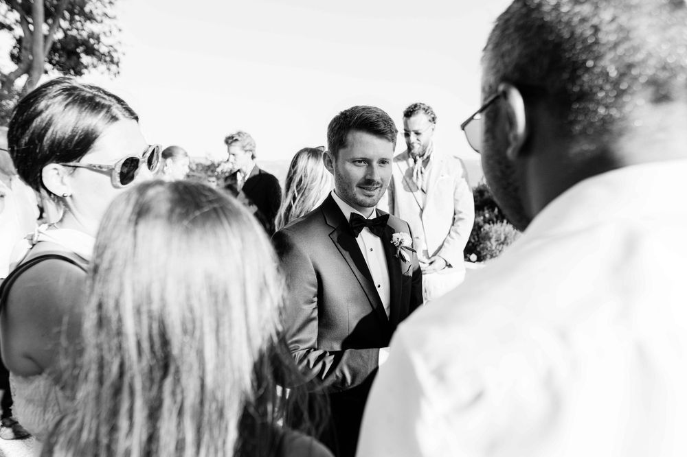 Groom in tuxedo greeting wedding guests outdoors in black and white photo.