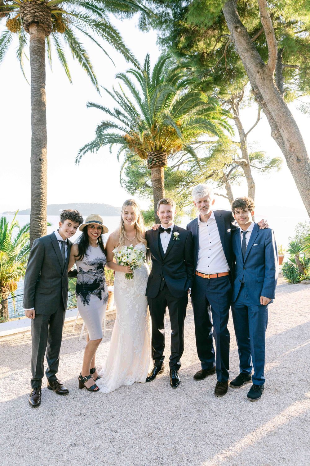 Wedding party posing outdoors near palm trees, bride in white gown with groom and guests in formal attire.