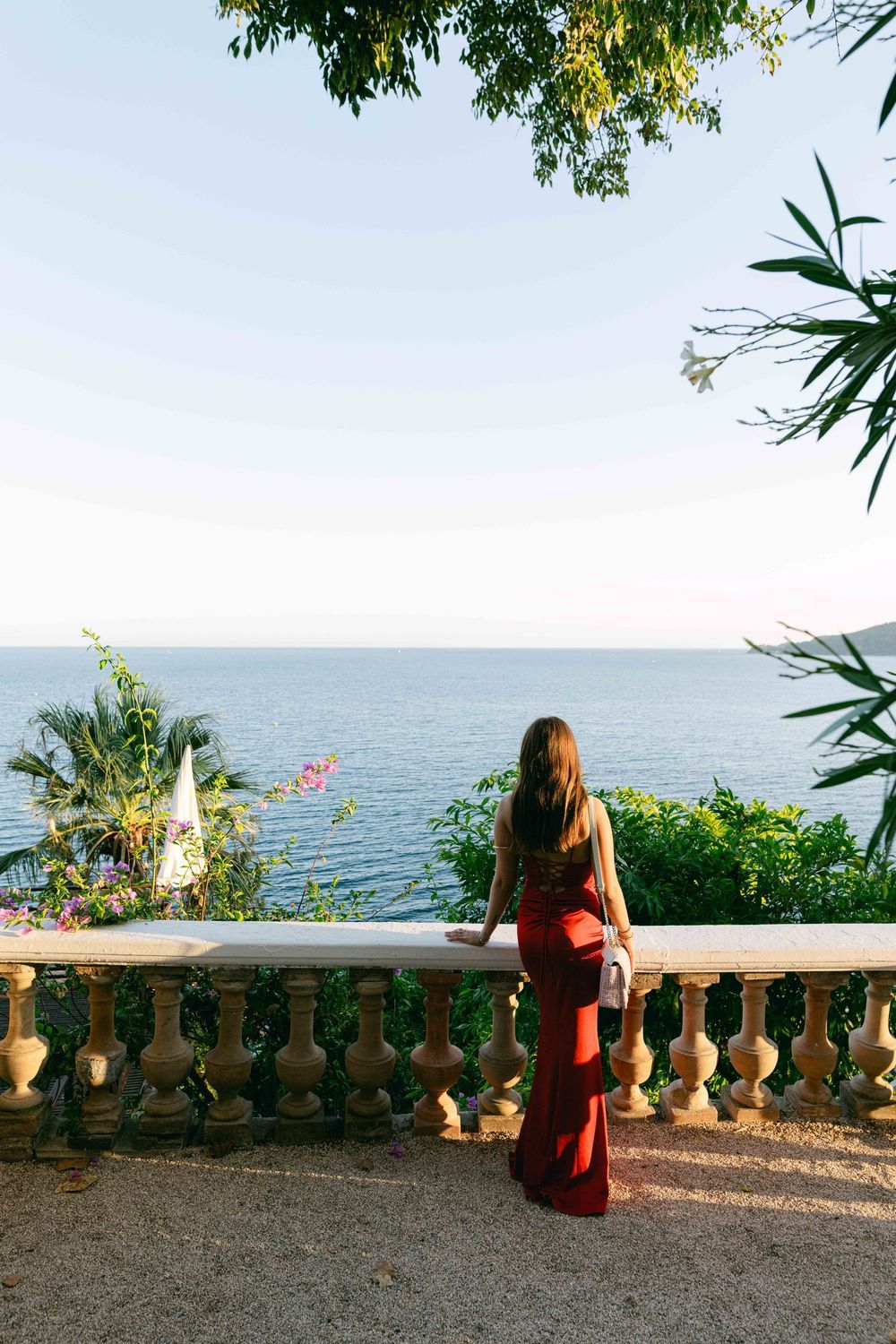 Woman in red gown stands on elegant stone terrace overlooking serene blue ocean, surrounded by lush tropical greenery.