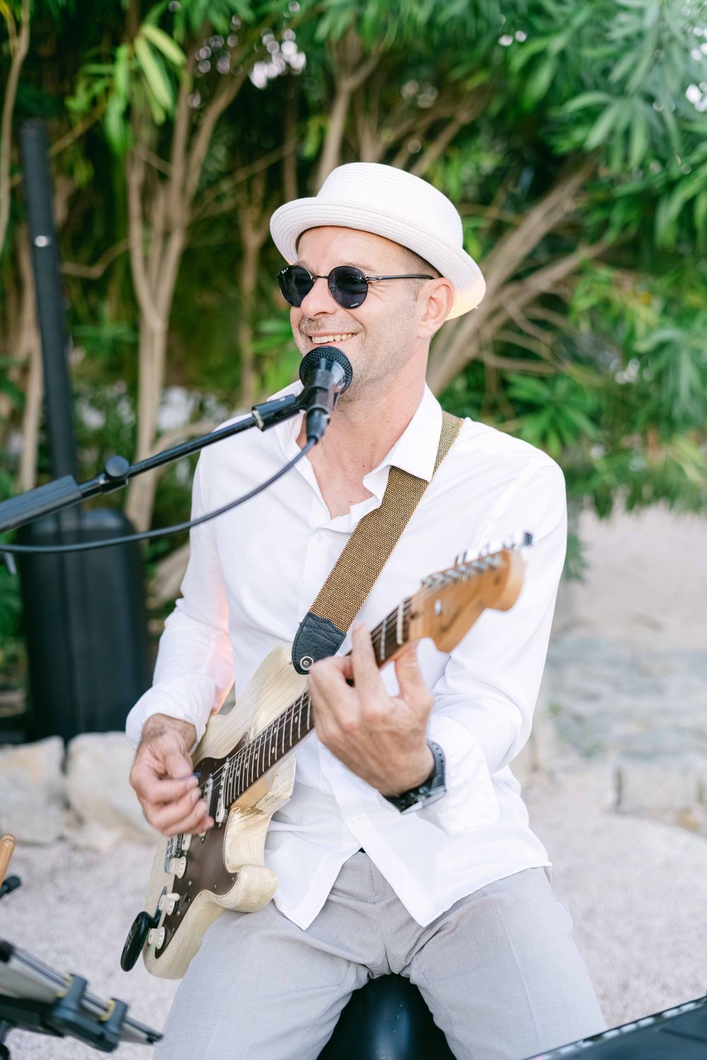 Smiling male musician in white hat and sunglasses playing electric guitar and singing into microphone outdoors.