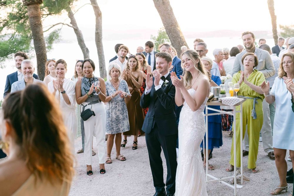 Bride and groom celebrated by clapping wedding guests at outdoor seaside reception.