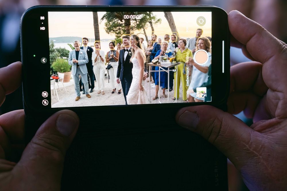 Hands holding a smartphone capturing a wedding couple kissing surrounded by cheering guests outdoors.