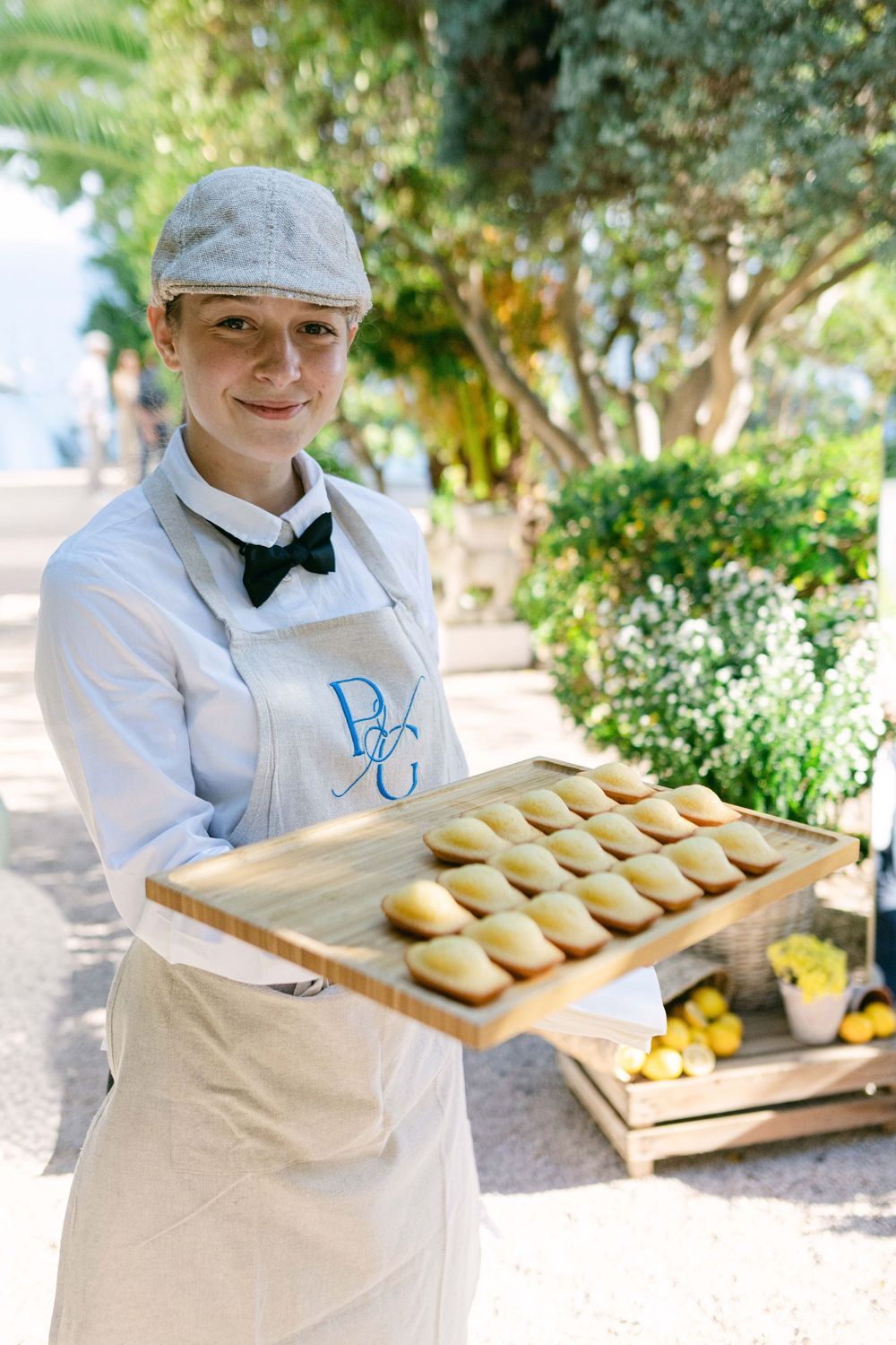 Smiling server in white apron and cap holds tray of madeleines outdoors at elegant garden event.