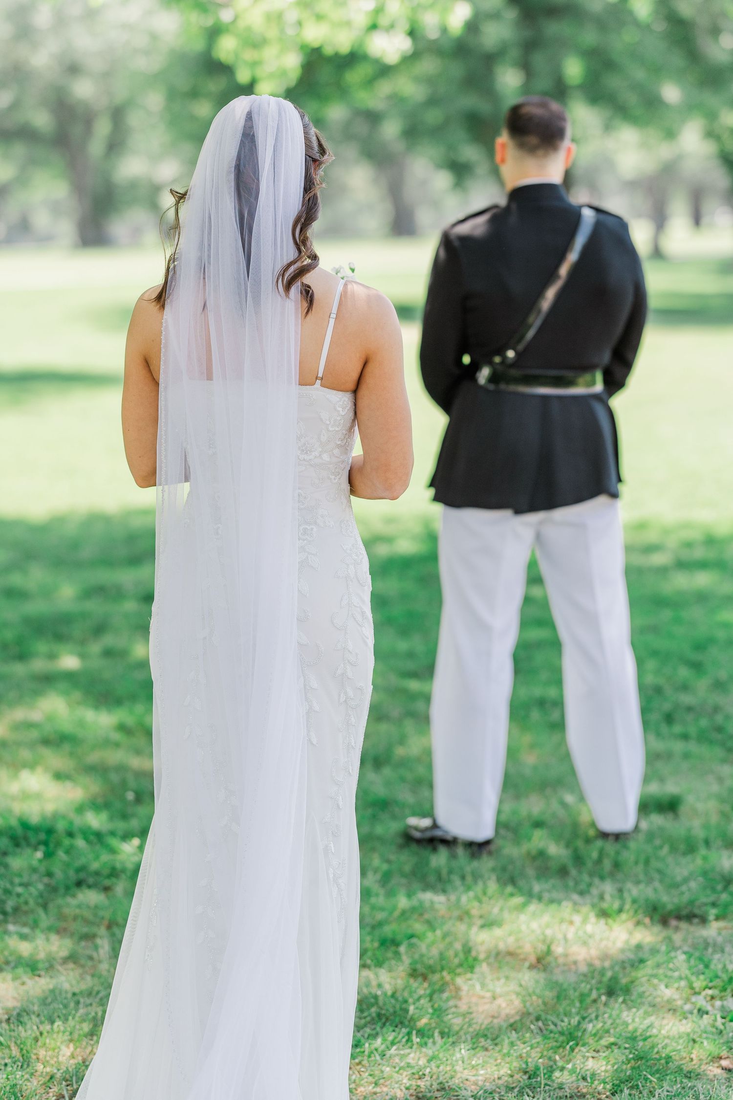 Bride in white gown and veil stands behind groom in military dress uniform during outdoor first look ceremony.