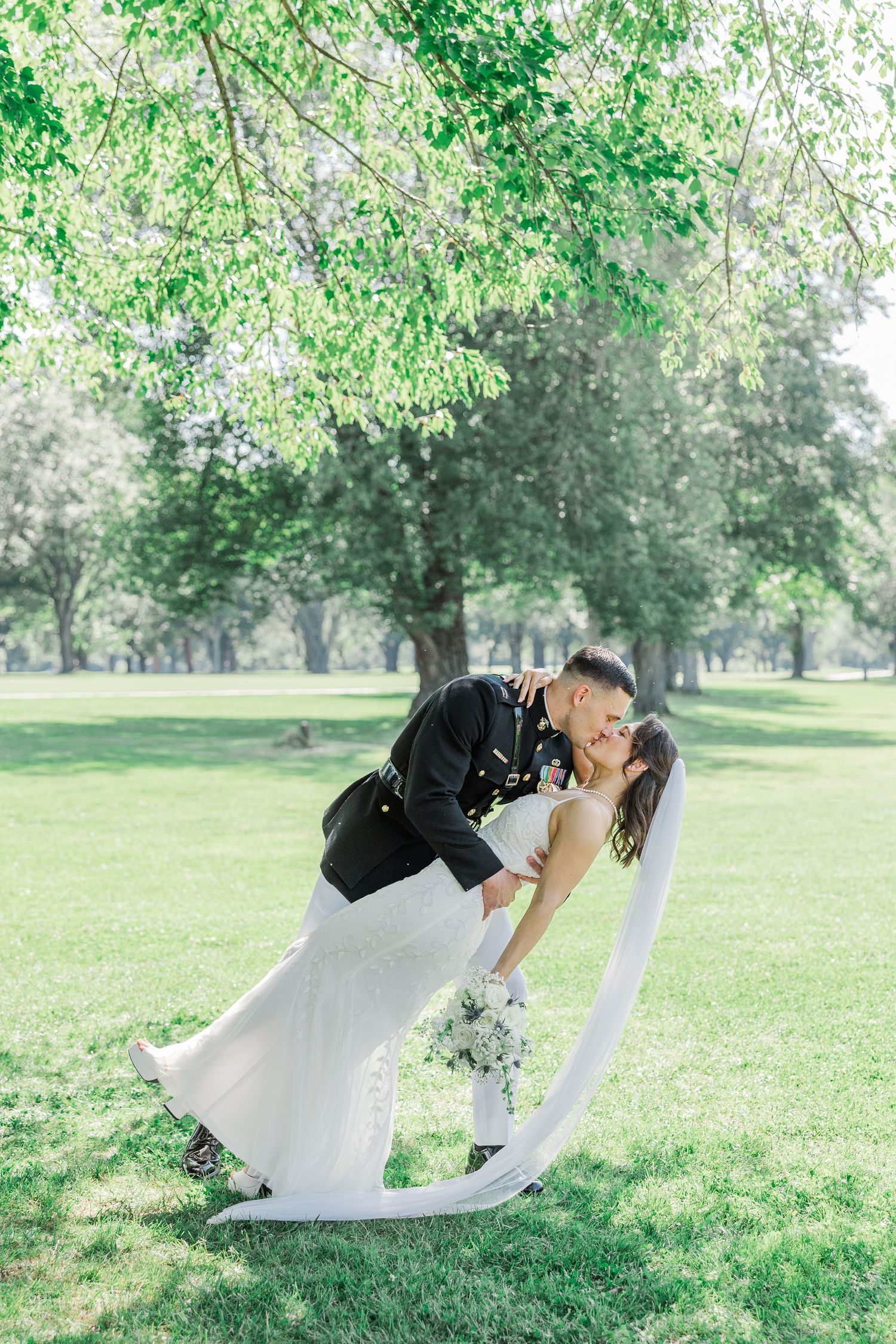 Military groom dips bride in white gown for romantic kiss outdoors in lush green park setting.