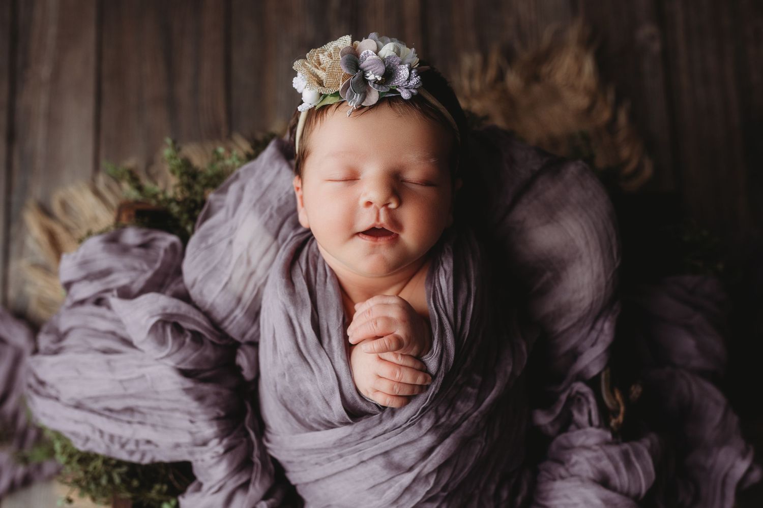 Sleeping newborn baby wrapped in purple cloth with floral headband in a cozy nest-like setting.
