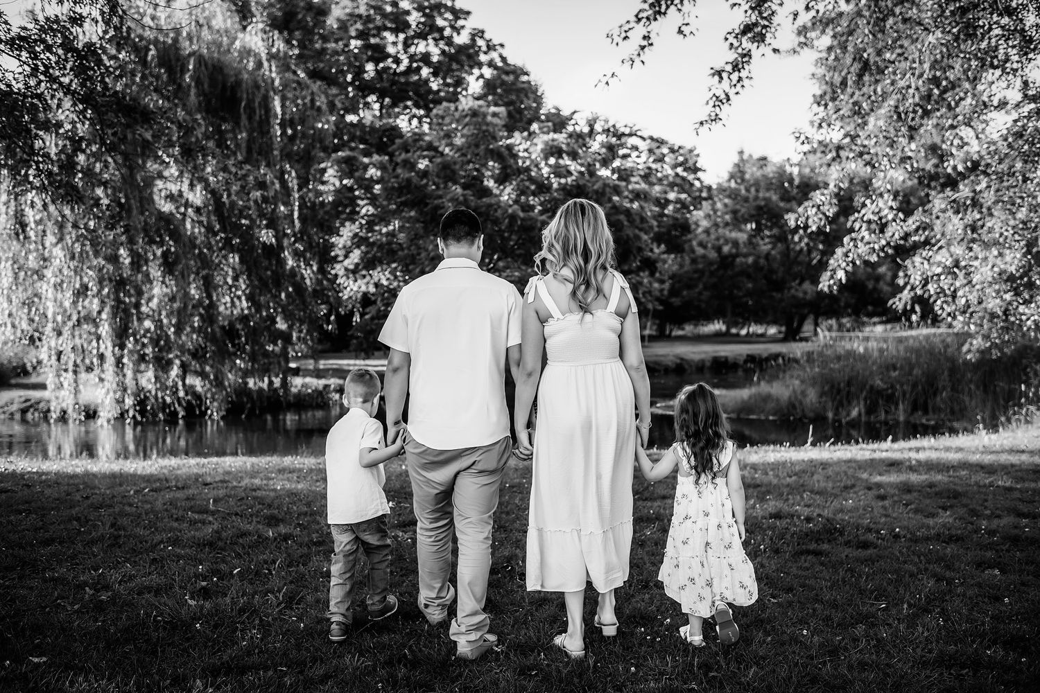 Black and white photo of a family of four holding hands while standing in a lush green park near a pond.