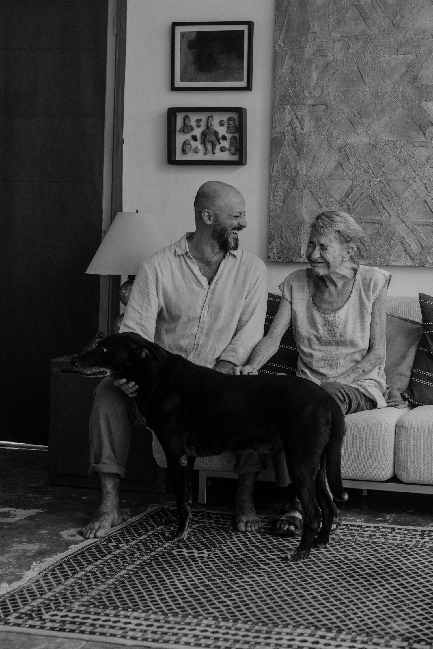 Black and white photo of a bald man and elderly woman smiling on a sofa with a black Labrador dog indoors.