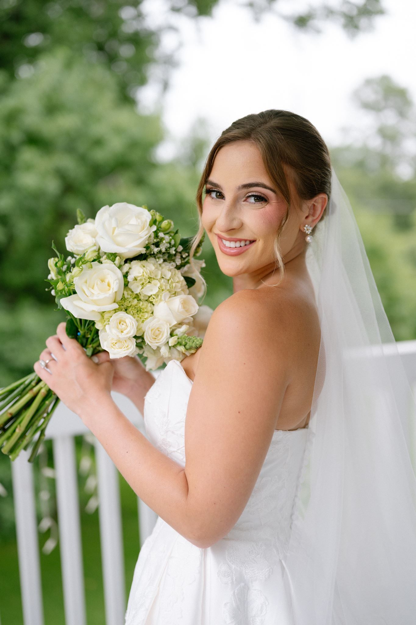 Smiling bride in strapless white gown holding elegant bouquet of white roses and green hydrangeas with veil outdoors.
