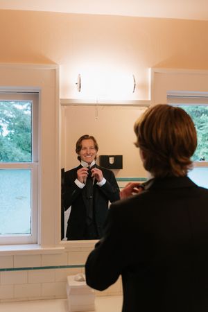 Groom smiling in bathroom mirror while adjusting his tie, dressed in a black suit on his wedding day.