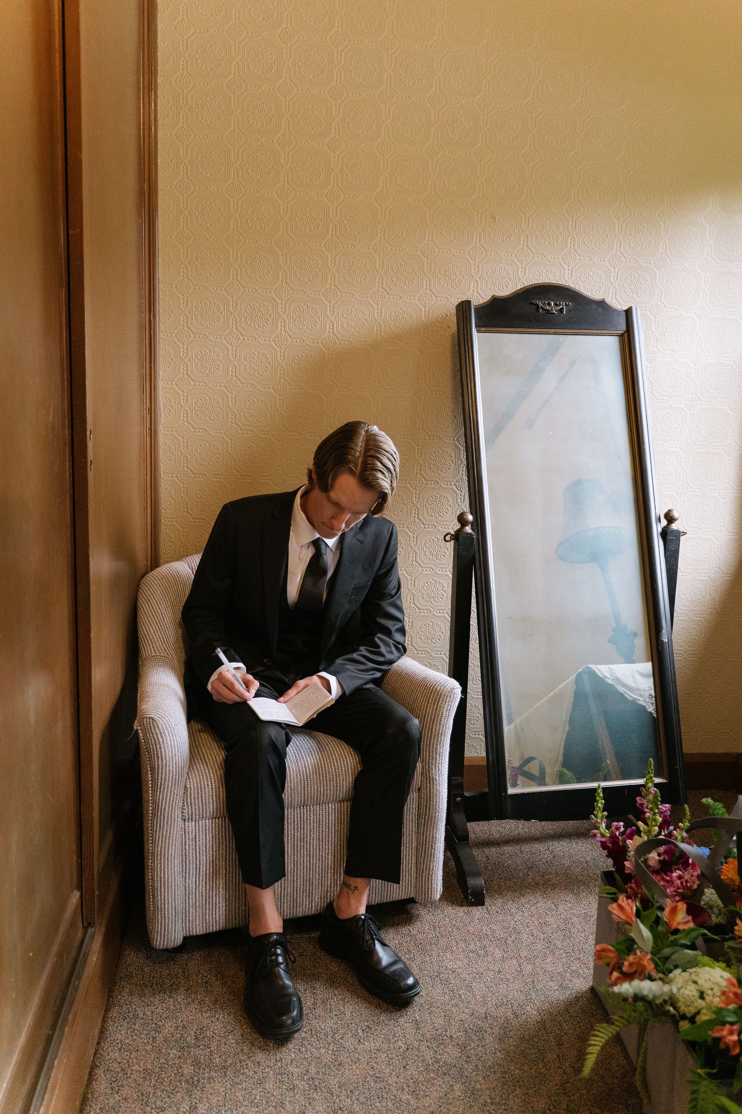 Groom in black suit writing vows while seated in a chair beside a floor mirror and wedding flowers.