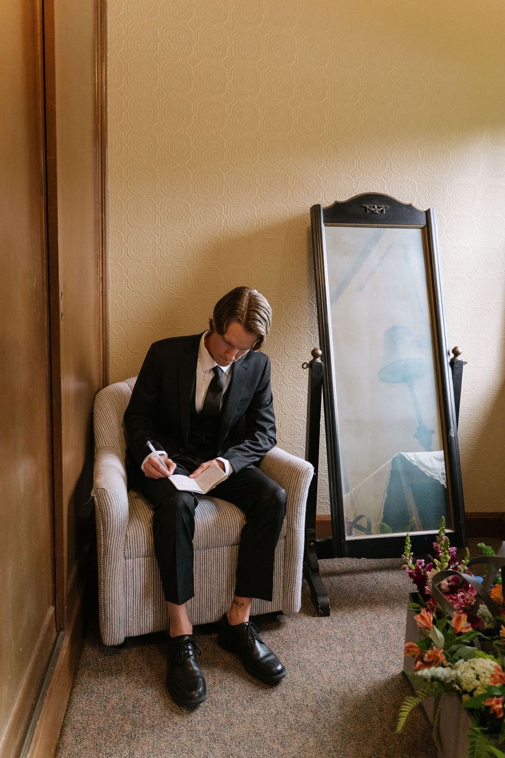 Groom in black suit writing vows while seated in a chair beside a floor mirror and wedding flowers.