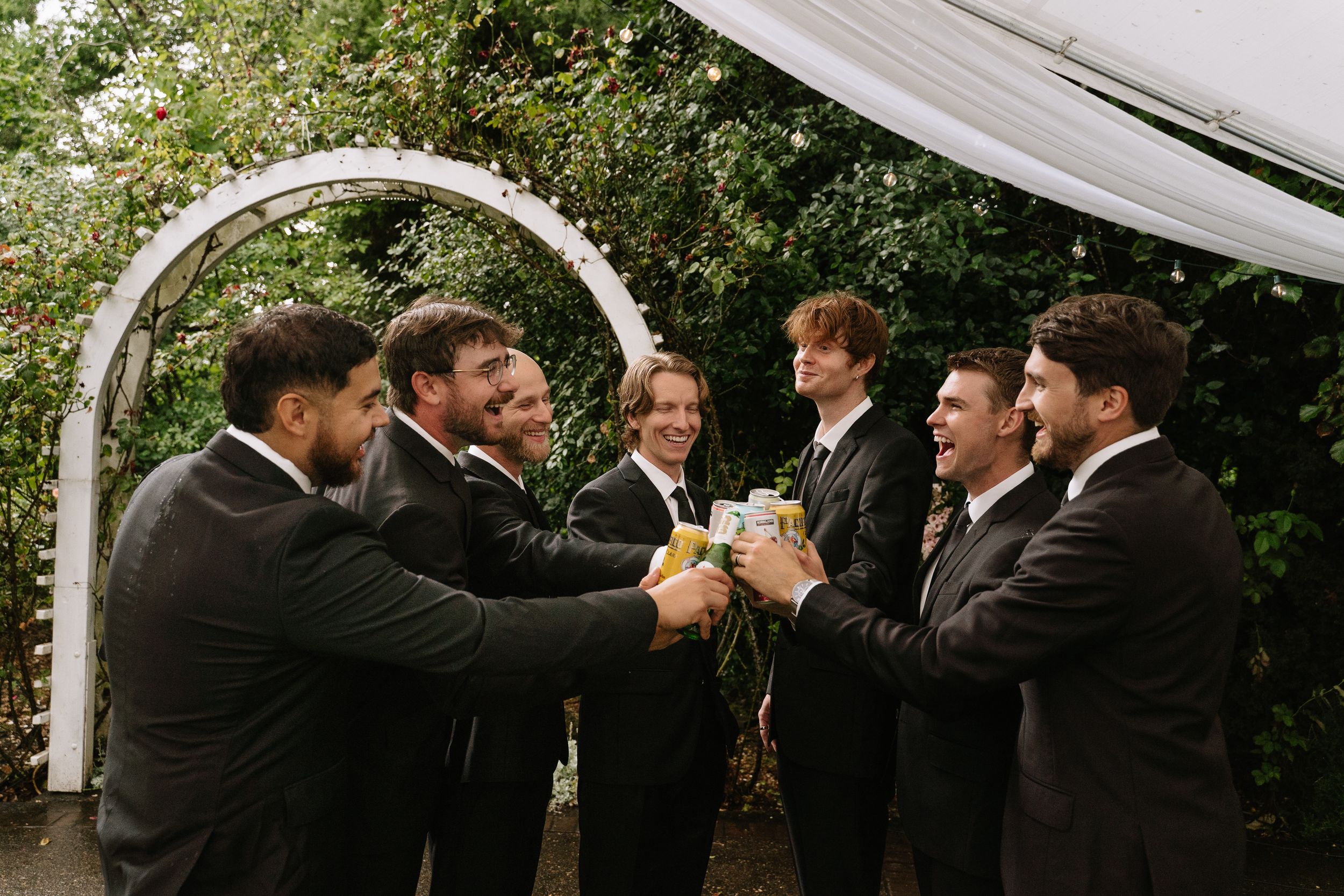 Groomsmen in black suits toasting drinks under a white arch at an outdoor wedding ceremony.