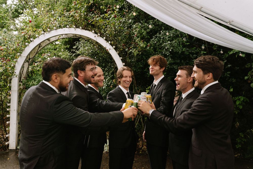 Groomsmen in black suits toasting drinks under a white arch at an outdoor wedding ceremony.