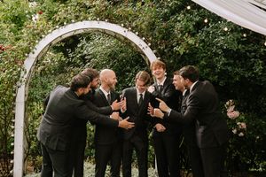 Groomsmen in black suits celebrate with the groom under a floral arch in an outdoor garden wedding setting.