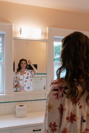 Woman in floral robe fixing her hair while looking at her reflection in a bathroom mirror.