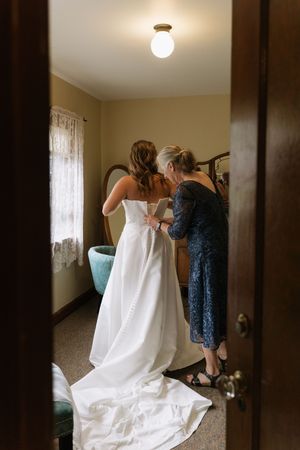 Bride in elegant white gown being helped with her dress by a woman in a blue lace outfit before wedding.