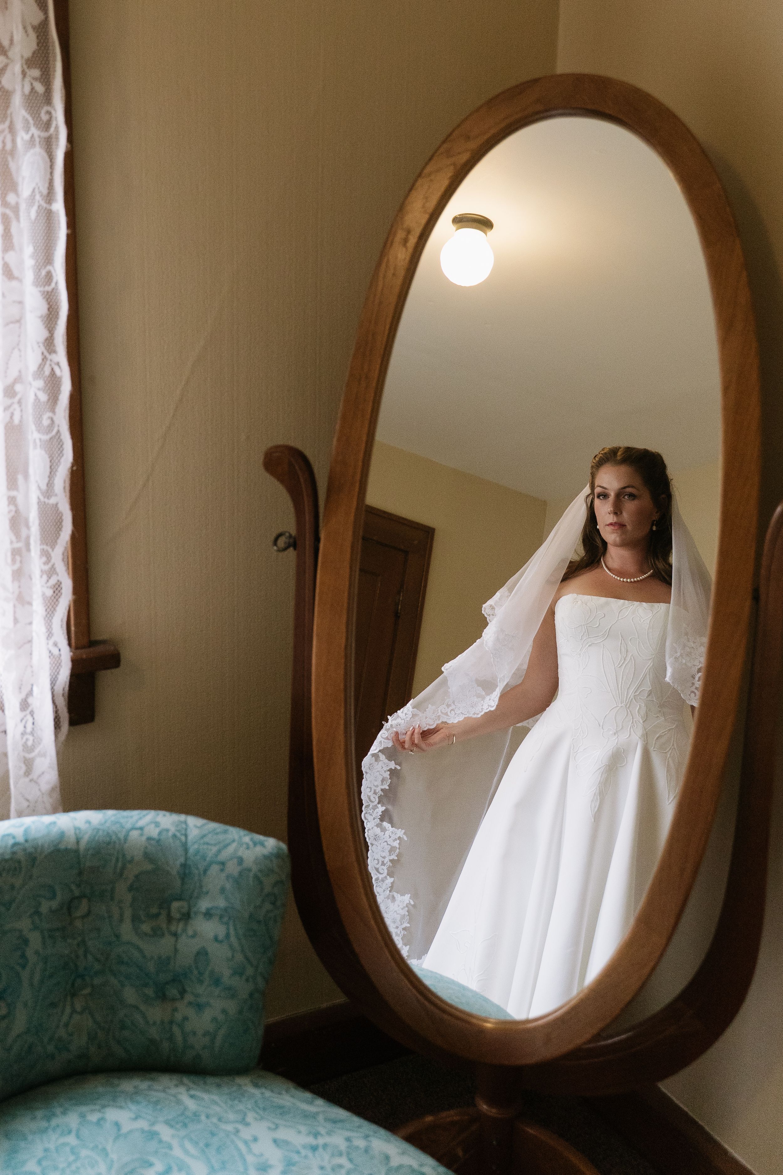 Bride in white wedding gown and veil reflected in ornate oval wooden mirror in vintage room with teal chair.