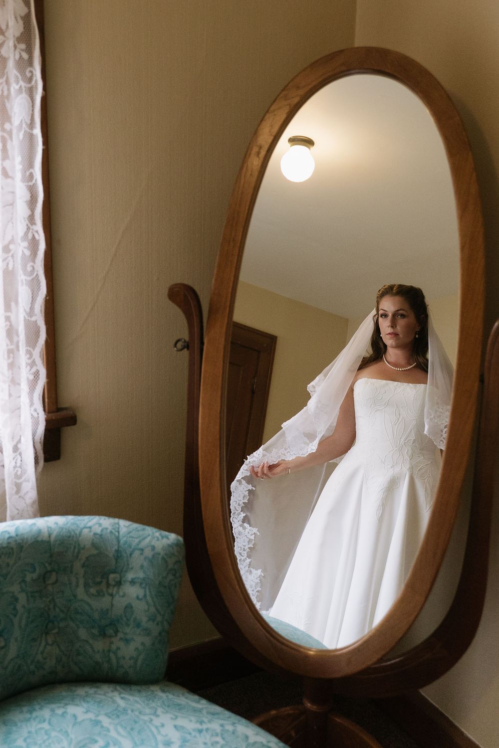 Bride in white wedding gown and veil reflected in ornate oval wooden mirror in vintage room with teal chair.