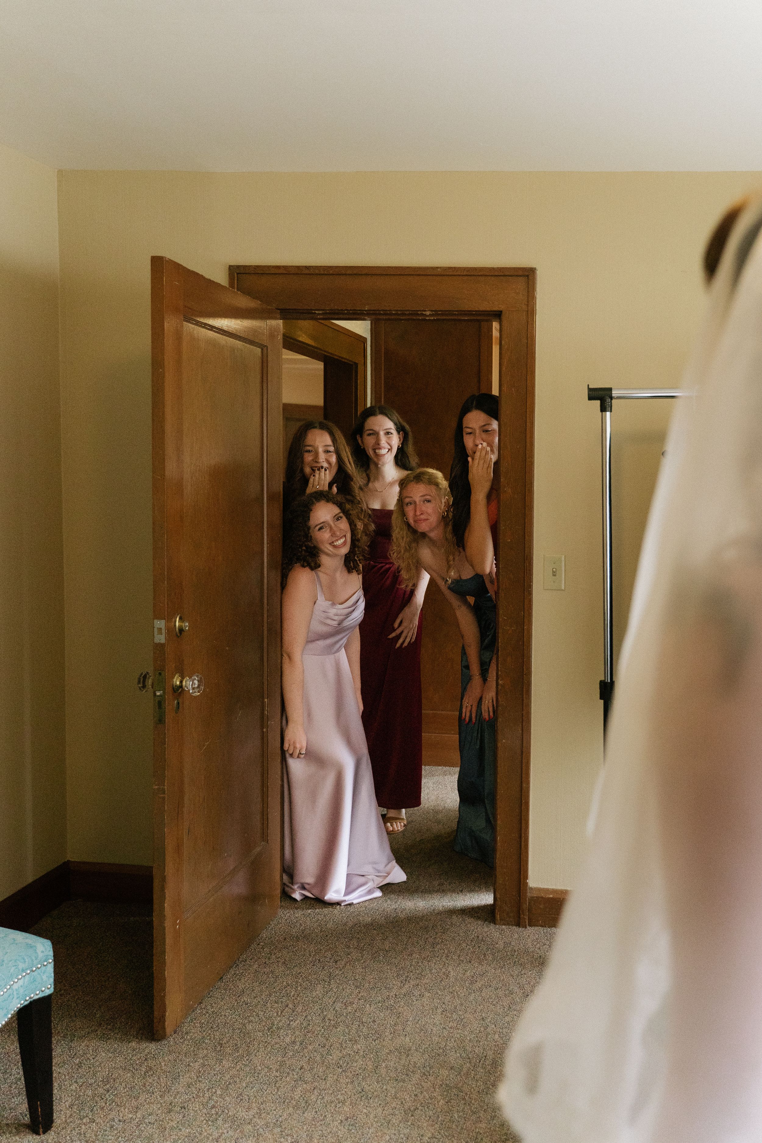 Bridesmaids peek through a doorway with joyful expressions as they see the bride in her wedding dress.