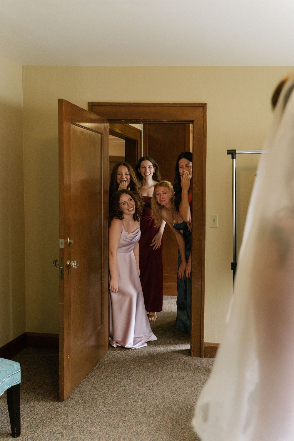Bridesmaids peek through a doorway with joyful expressions as they see the bride in her wedding dress.