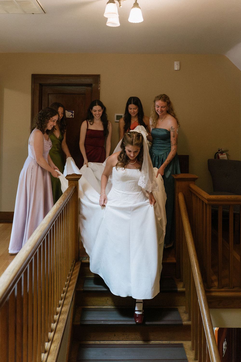 Bride descends staircase in white gown assisted by bridesmaids in colorful dresses during wedding preparations.