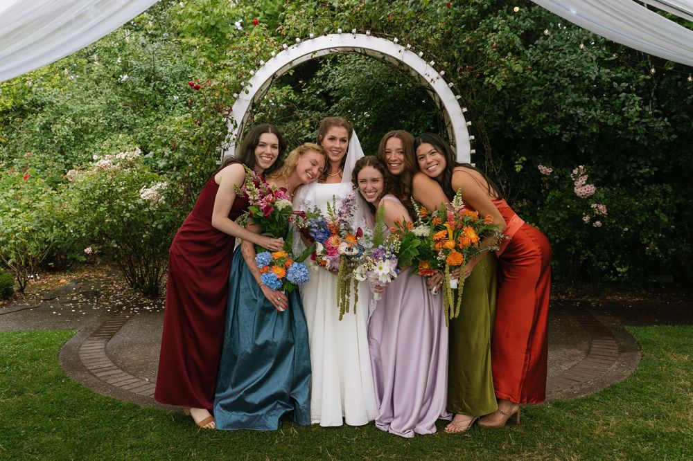 Bride in white gown poses with five bridesmaids in colorful dresses holding bouquets under a garden arch.