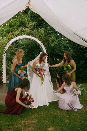 Bride in white gown surrounded by bridesmaids in colorful dresses under a floral arch at an outdoor wedding.
