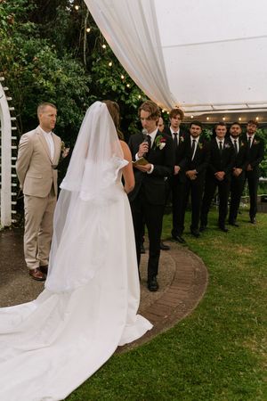 Outdoor wedding ceremony with bride in white gown and veil facing groom, groomsmen in black suits lined up on green lawn.