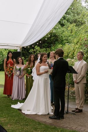Outdoor wedding ceremony with bride in white gown, colorful bridesmaids, and groom under white tent canopy.