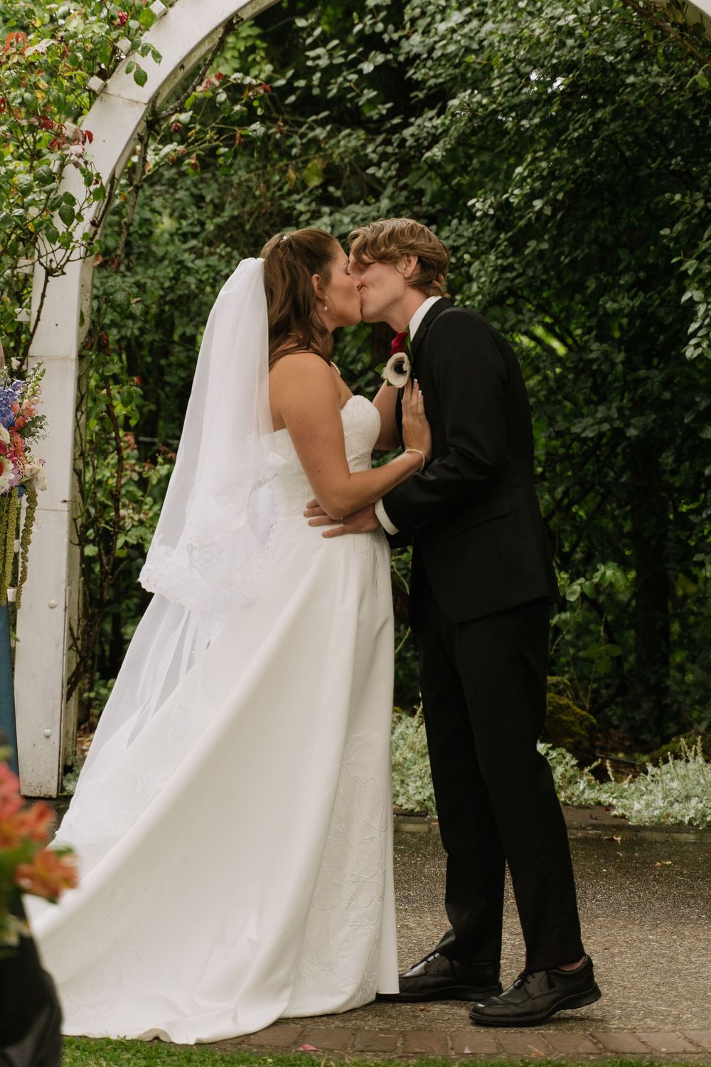 Bride and groom share first kiss under floral arch, bride in white gown with veil, groom in black suit.