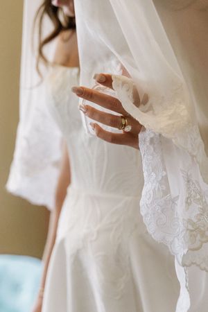 Bride in lace wedding dress holding sheer veil, showcasing elegant gold diamond engagement ring and manicured nails.