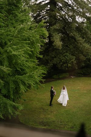 Bride and groom share a romantic moment on lush green lawn surrounded by tall trees on wedding day.