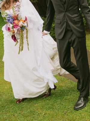 Bride in white lace gown holding colorful bouquet walks with groom in dark suit on green grass.