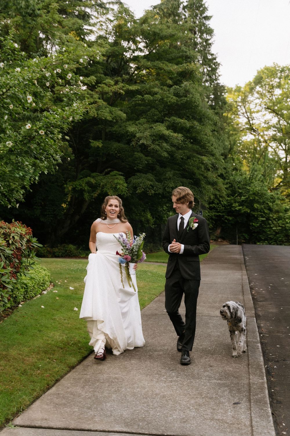 Bride and groom walk their dog along a garden path, bride holding wildflower bouquet in white gown.