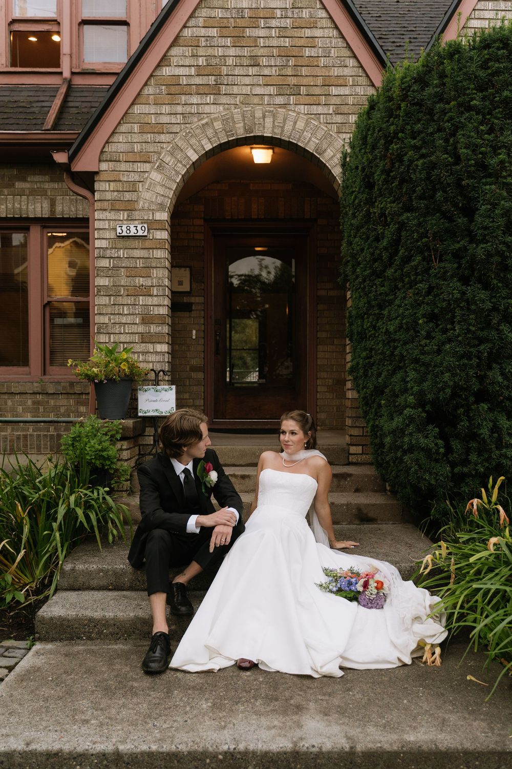 Bride and groom sit together on steps of brick building with arched doorway holding colorful bouquet.
