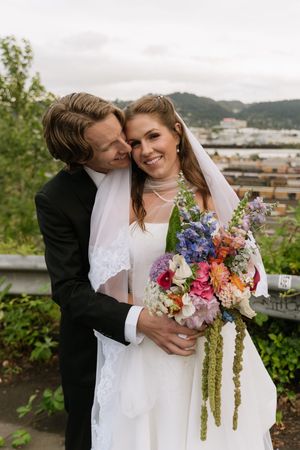 Groom embraces bride from behind as she holds vibrant wildflower bouquet with scenic hills in background.