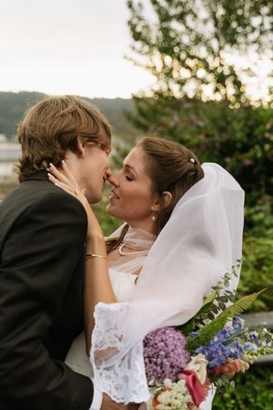 Bride and groom share a kiss outdoors with lace veil and colorful floral bouquet at golden hour.
