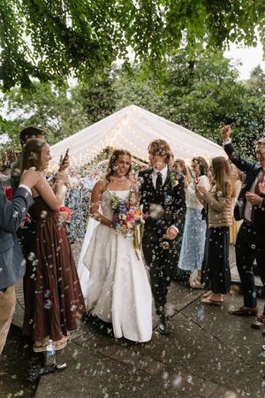 Bride and groom walk through confetti and bubbles as smiling wedding guests celebrate their exit under a marquee.