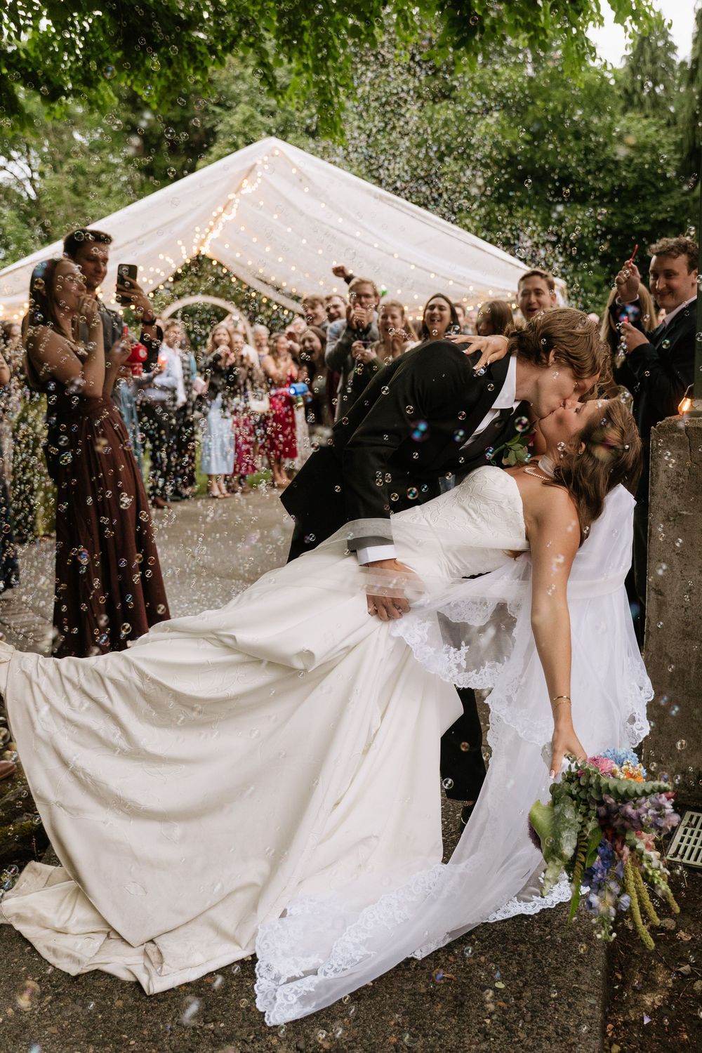 Groom dips bride for a romantic kiss as wedding guests celebrate with confetti outdoors.