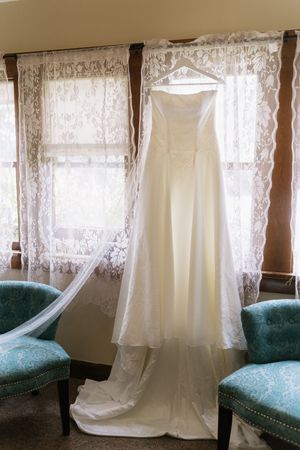 Elegant strapless white wedding gown with lace veil hanging in window between two teal accent chairs.