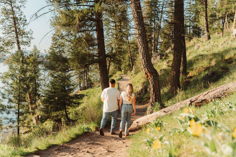 A couple walks hand-in-hand along a scenic forest trail surrounded by pine trees and wildflowers near a lake.