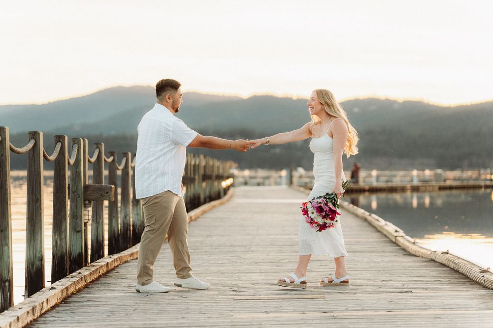 Couple holding hands on wooden pier at sunset, woman in white dress holding bouquet, mountains reflecting on water.