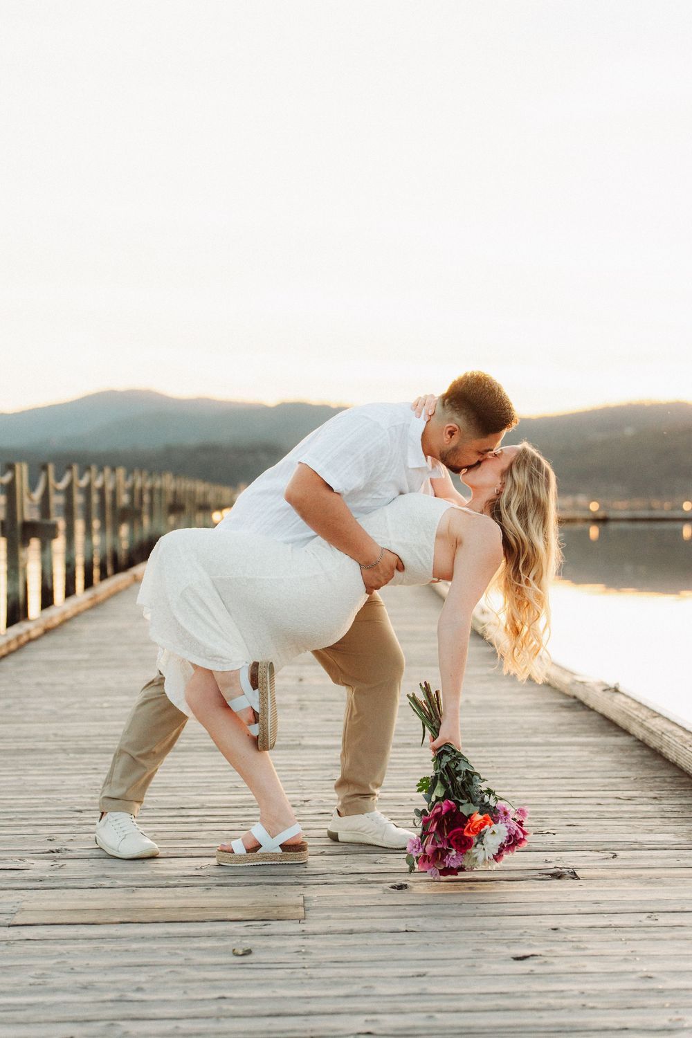 Couple sharing a romantic dip kiss on a wooden pier at sunset with flowers.