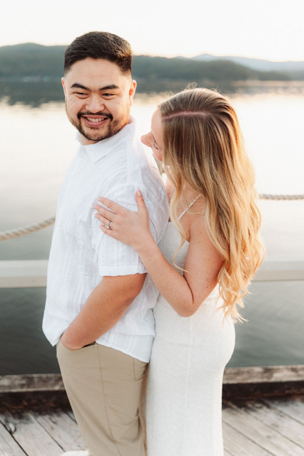 Happy couple embracing on a dock at sunset with a lake and mountains in the background.