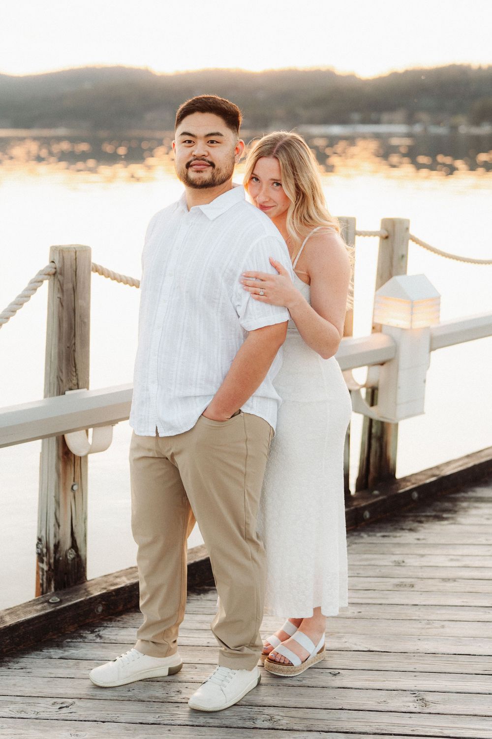 Engaged couple posing on a wooden dock at sunset, man in white shirt and woman in white dress.
