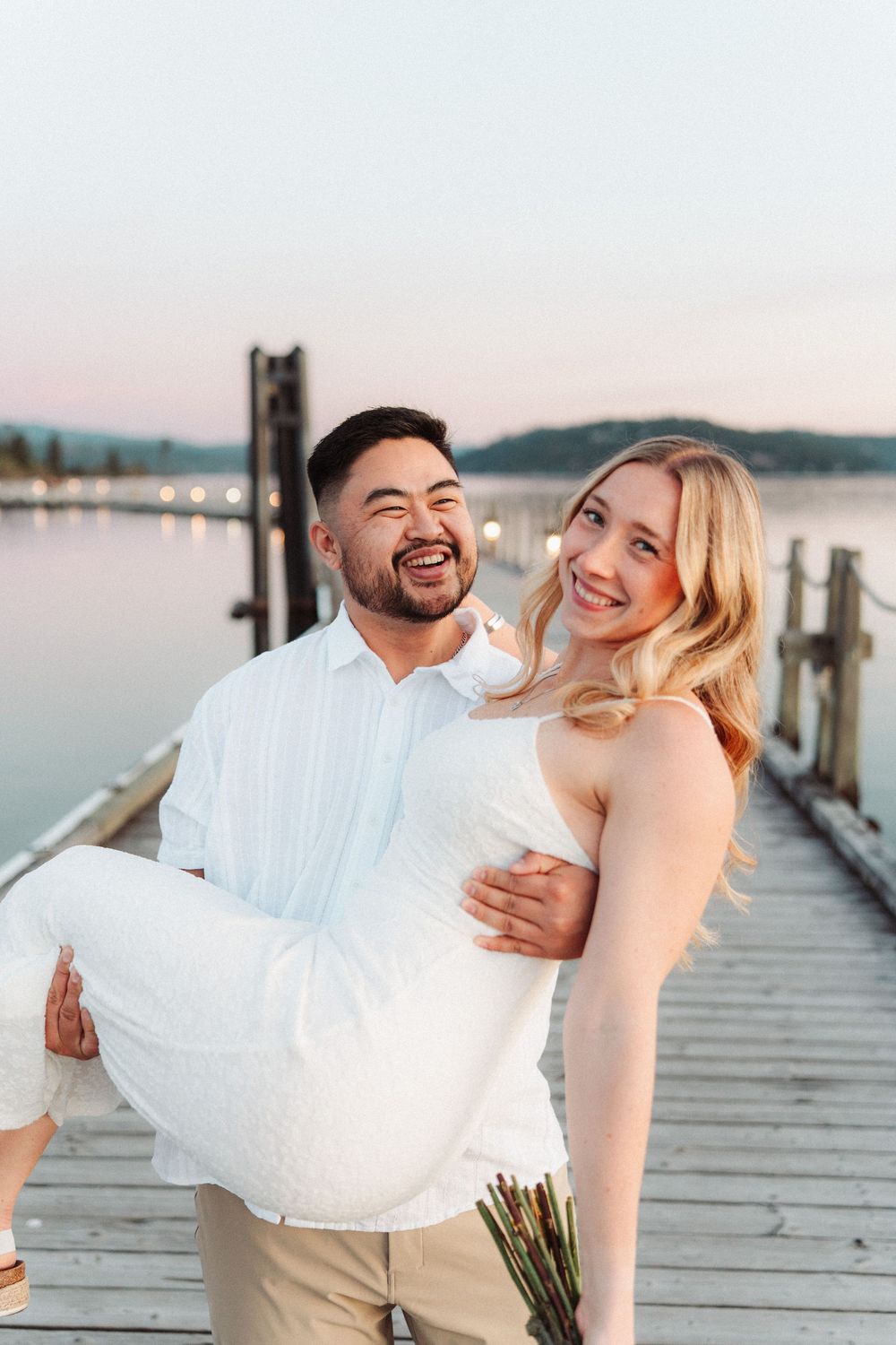 Joyful man lifting his smiling fiancee on a waterfront dock during a romantic engagement session.