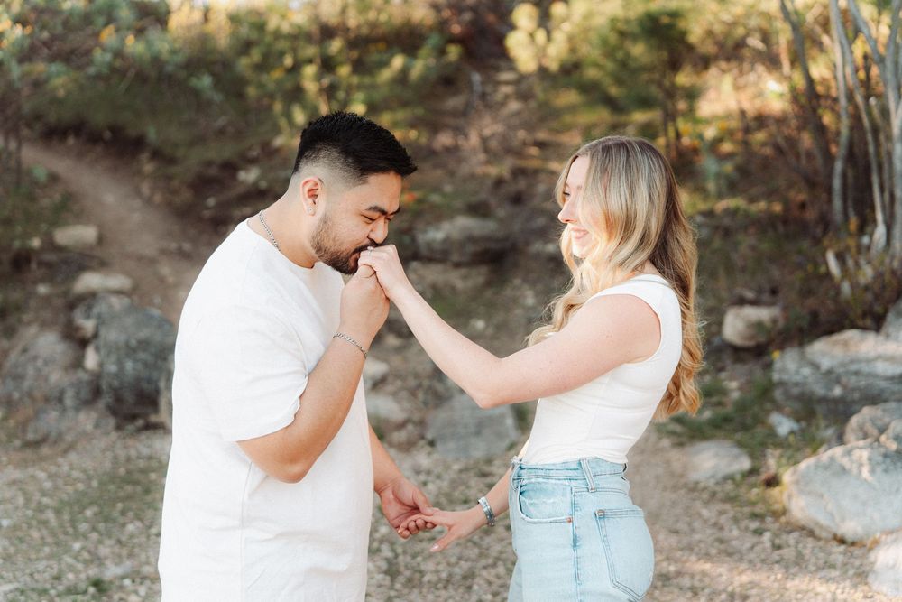 Man gently kissing womans hand outdoors, both wearing white tops, surrounded by rocky natural landscape in warm light.