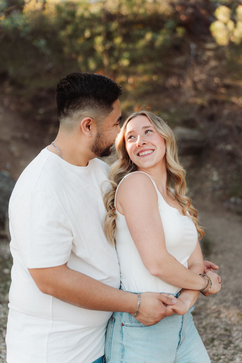 Romantic couple embracing outdoors, man whispering to smiling blonde woman in white tank top and jeans at sunset.